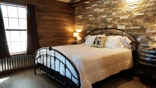 A bedroom with a stone wall, a bed with a metal frame, a window with curtains, and a nightstand with a lamp. - Bakery Haus- Tres Leches | Pleasant Cabin for 2 (Fredericksburg)
