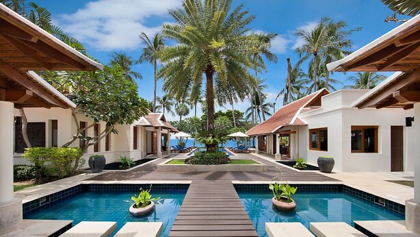 A resort pool with a wooden walkway, palm trees, and white buildings with red roofs. - Baan Chao Lay (Koh Samui)