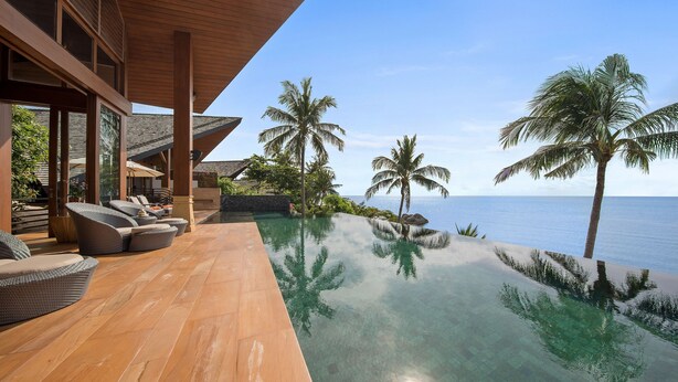 Une piscine à débordement moderne, avec une eau cristalline qui reflète les palmiers et l’océan, une terrasse en bois avec des sièges et une vue imprenable sur la mer.