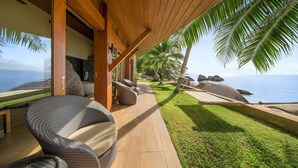 Une terrasse en bois avec des meubles en osier, une vue sur l’océan et une pelouse bien entretenue.