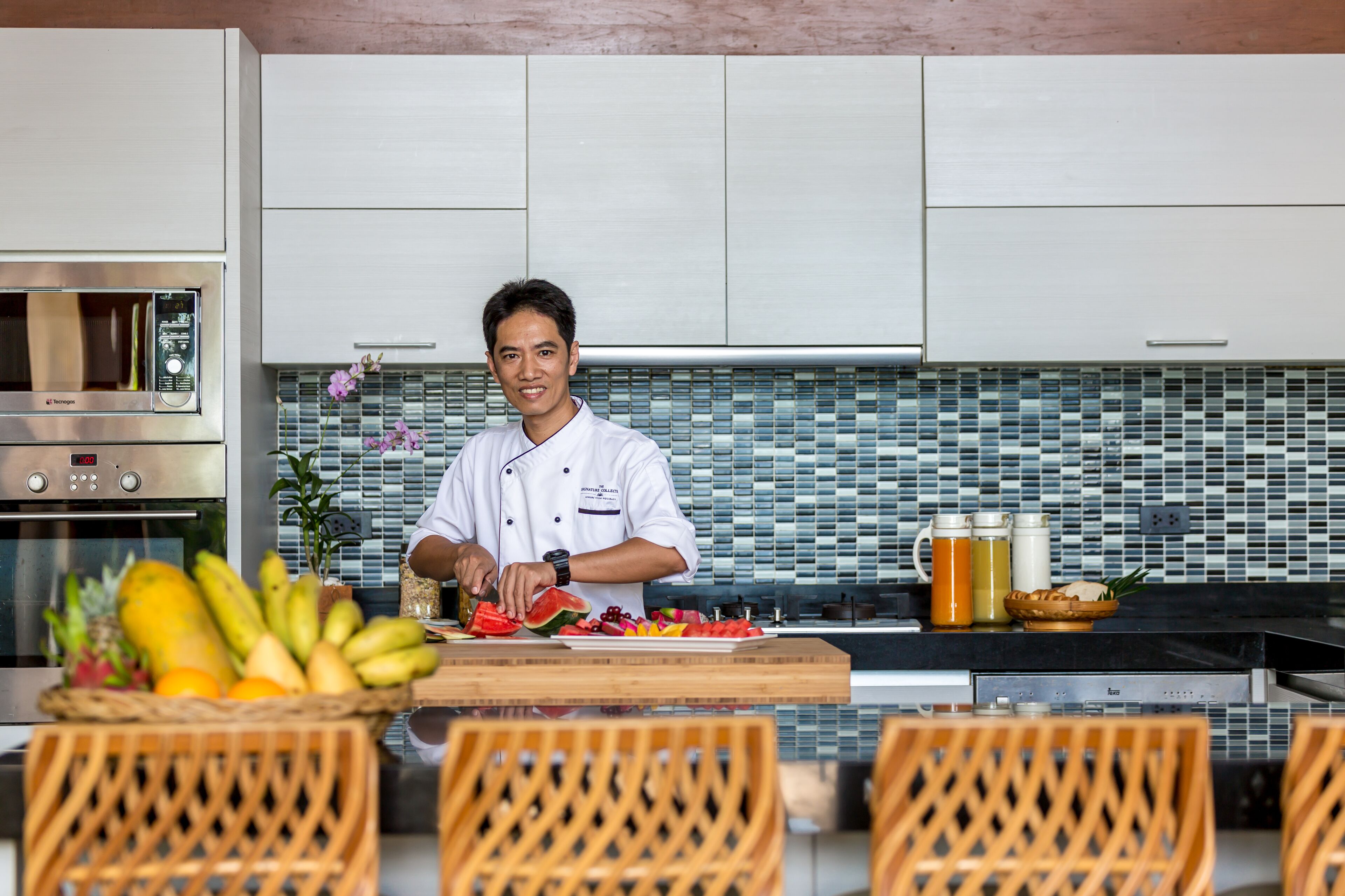 A chef in a modern kitchen with a tiled backsplash, stainless steel appliances, and a wooden dining table with chairs.