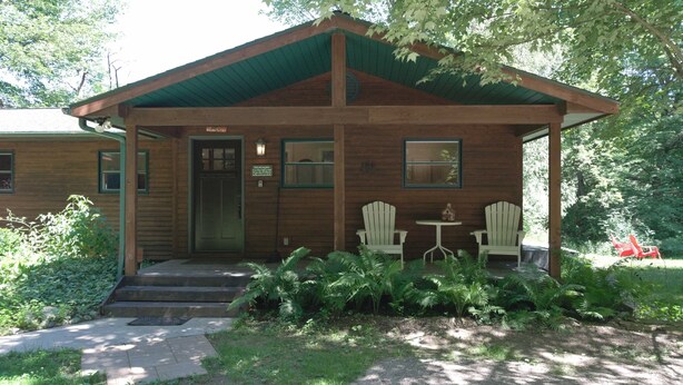 Eine Holzhütte mit Veranda, zwei Adirondack-Stühlen und einem kleinen Tisch.