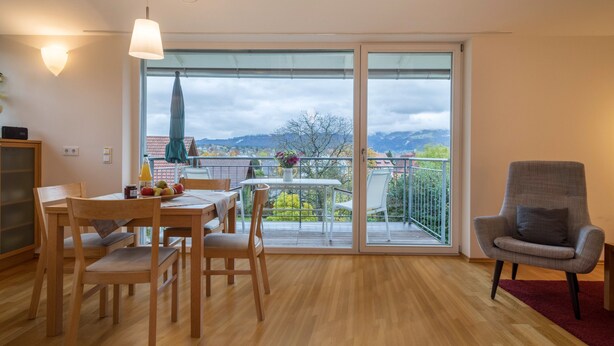 A dining area with a table and chairs, a balcony with a view of mountains, and a chair with a cushion. - Apartment ‘Pfirsichblüte’ on idyllic Family Farm House with Balcony & Mountain View (Lindau)