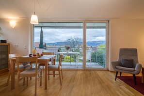 A dining area with a table and chairs, a balcony with a view of mountains, and a chair with a cushion. - Apartment ‘Pfirsichblüte’ on idyllic Family Farm House with Balcony & Mountain View (Lindau)