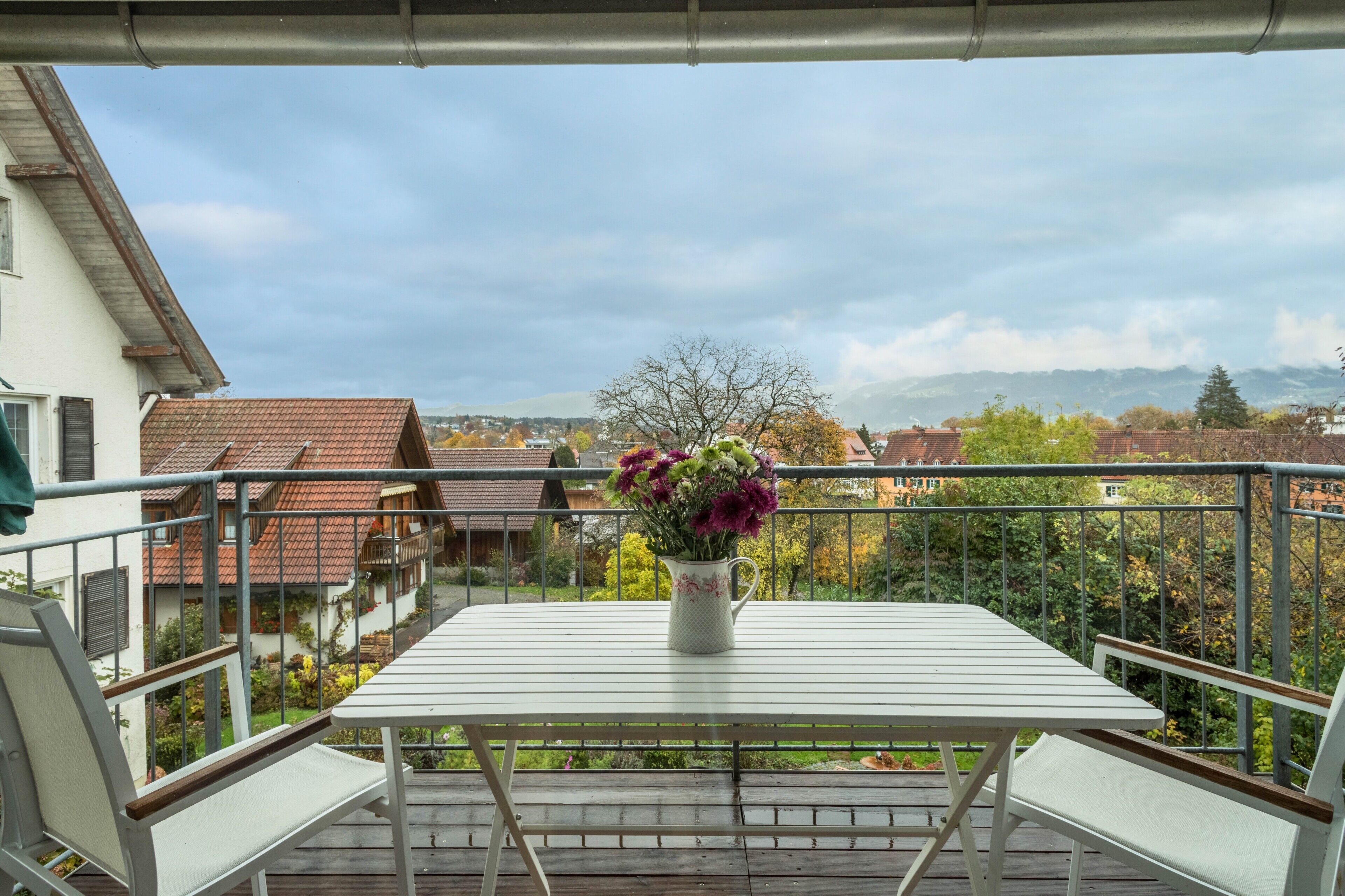 Ein Balkon mit Tisch und Stühlen, einer Blumenvase und Blick auf Häuser und Bäume.