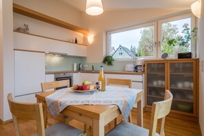 A kitchen with a dining table, chairs, a fruit bowl, a bottle, and a window with a view of houses.