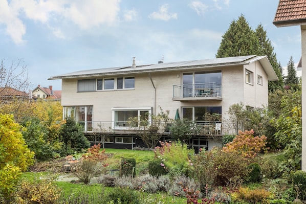 A two-story residential building with a balcony, surrounded by a garden with various plants and shrubs.
