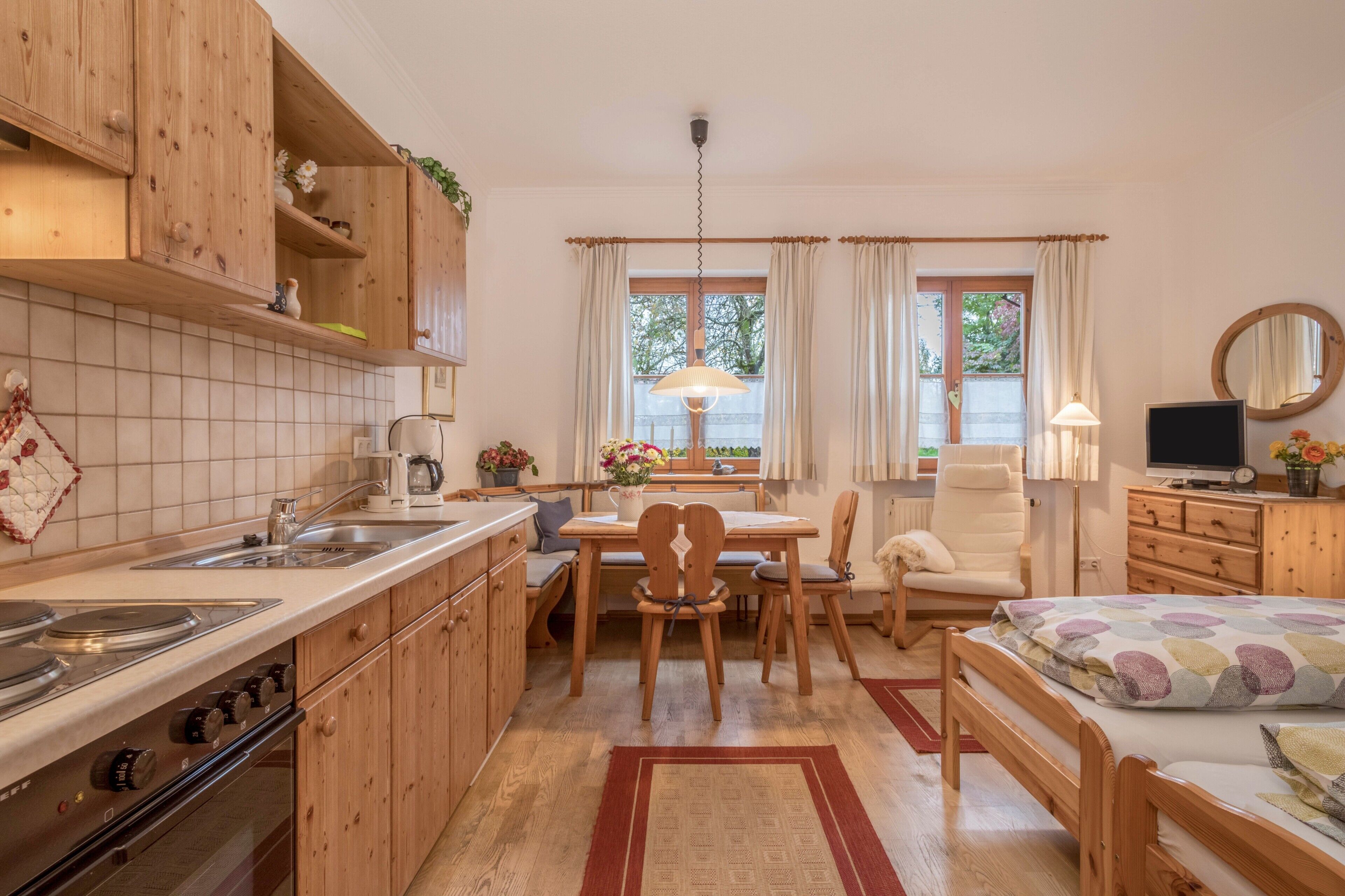 Eine kleine Küche mit Holzschränken, ein Essbereich mit Tisch und Stühlen, ein Bett mit gemustertem Überwurf und ein Fernseher auf einer Kommode.