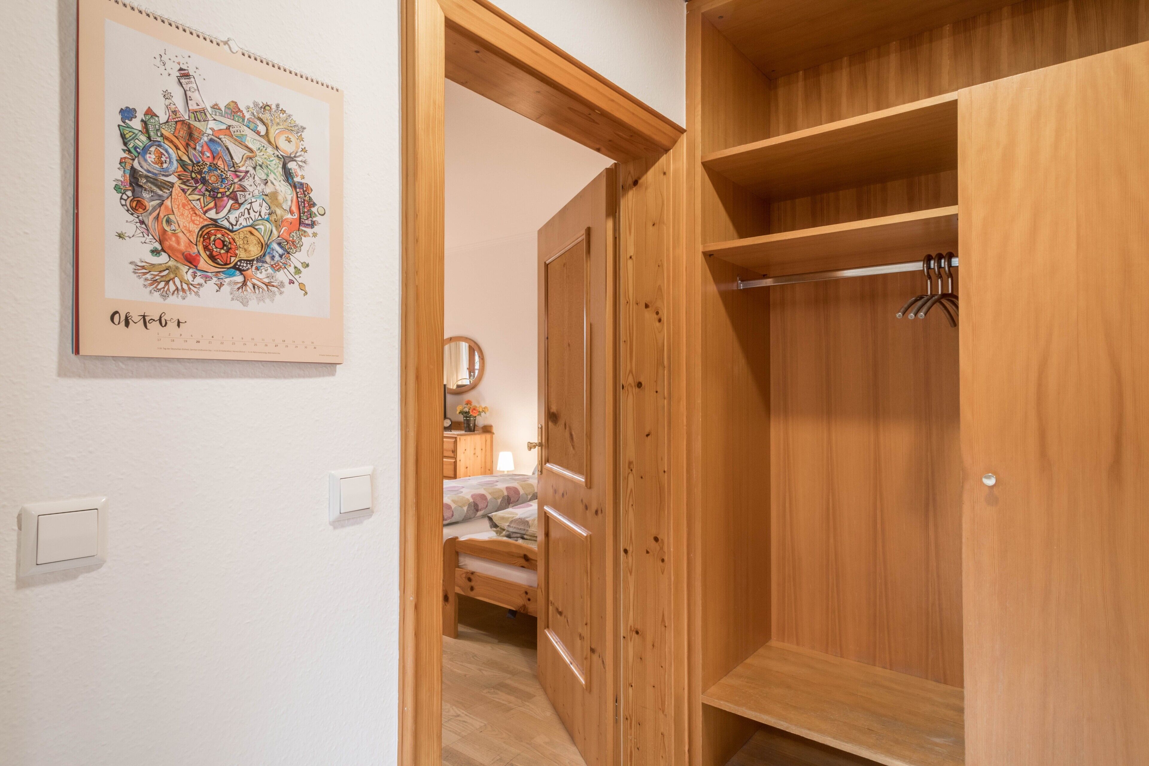 Ein Holzkleiderschrank mit Kleiderstange, ein Bett mit floraler Bettdecke und ein an der Wand befestigter Bilderrahmen.