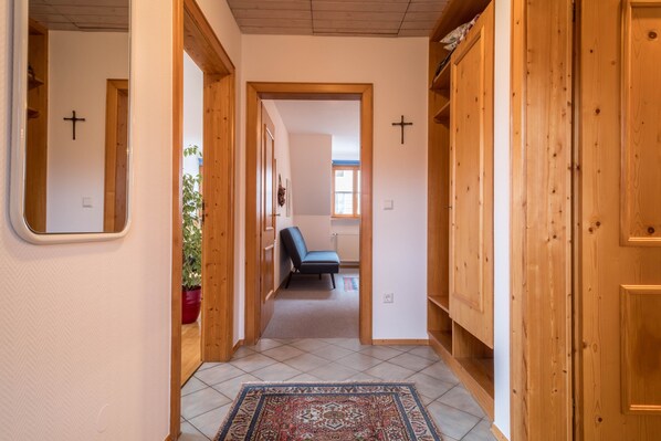 En hall med trädörrar, ett kors på väggen, en stol och en krukväxt.
