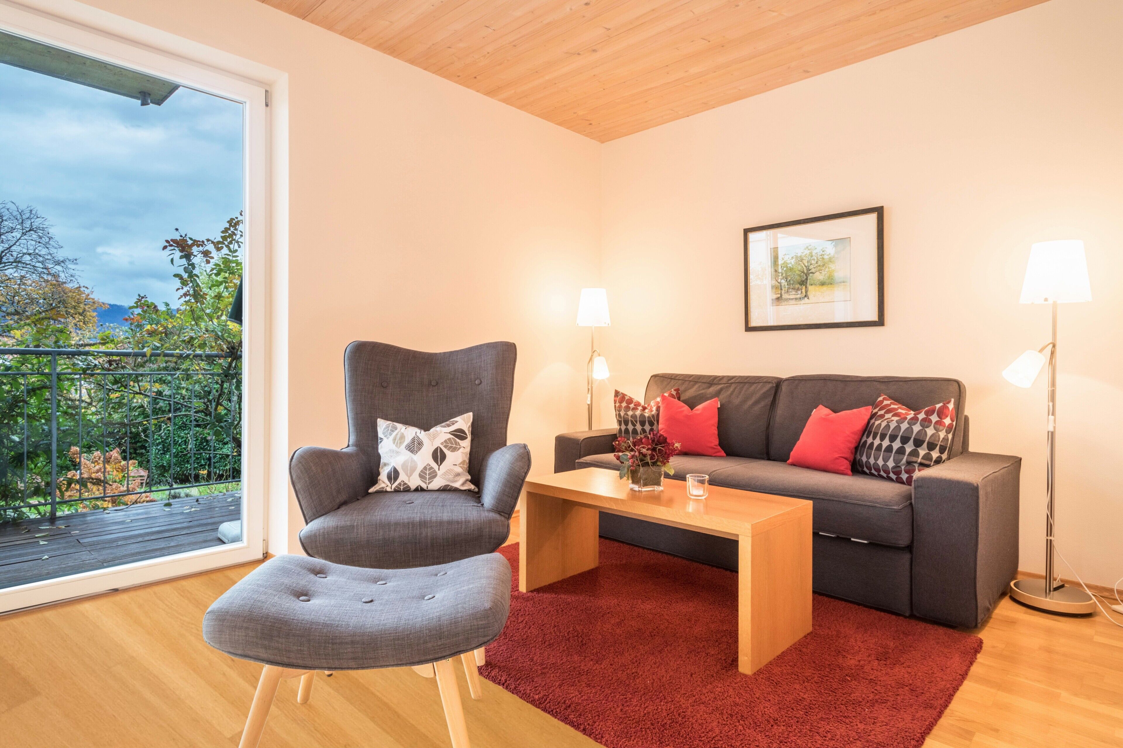 Un salon moderne avec un fauteuil gris, un canapé agrémenté de coussins rouges et à motifs, une table basse en bois et une grande fenêtre donnant sur un paysage verdoyant.