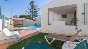 Zona de piscina exterior moderna amb gandules i una taula petita, envoltada per un prat ben cuidat i un edifici de pedra amb fonaments blancs.