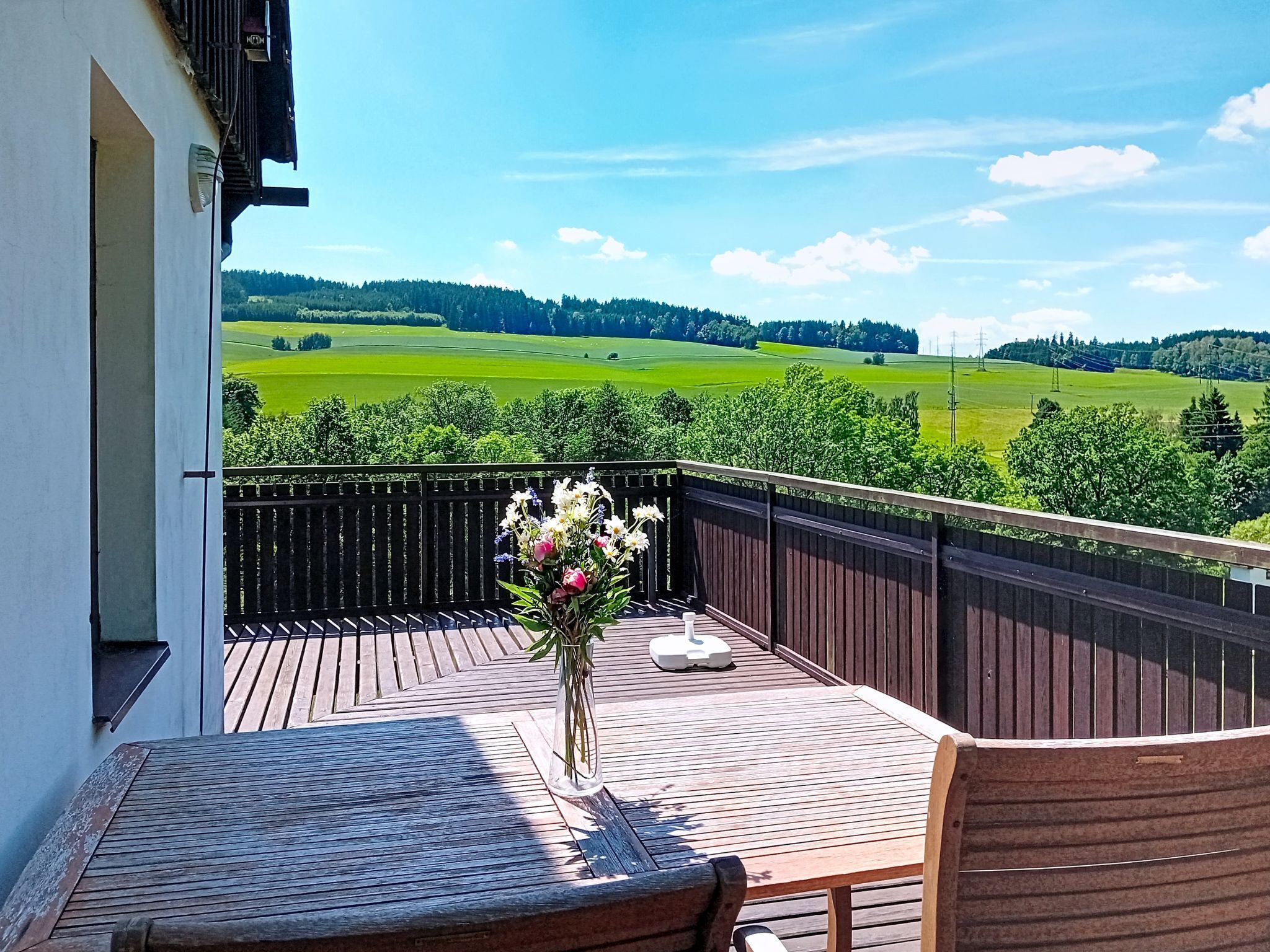 Eine Holzterrasse mit Tisch und Stühlen, eine Vase mit Blumen und Blick auf grüne Felder und Bäume.