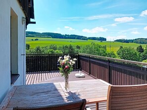 Una terraza de madera con una mesa y sillas, un florero con flores, y vistas a campos verdes y árboles.