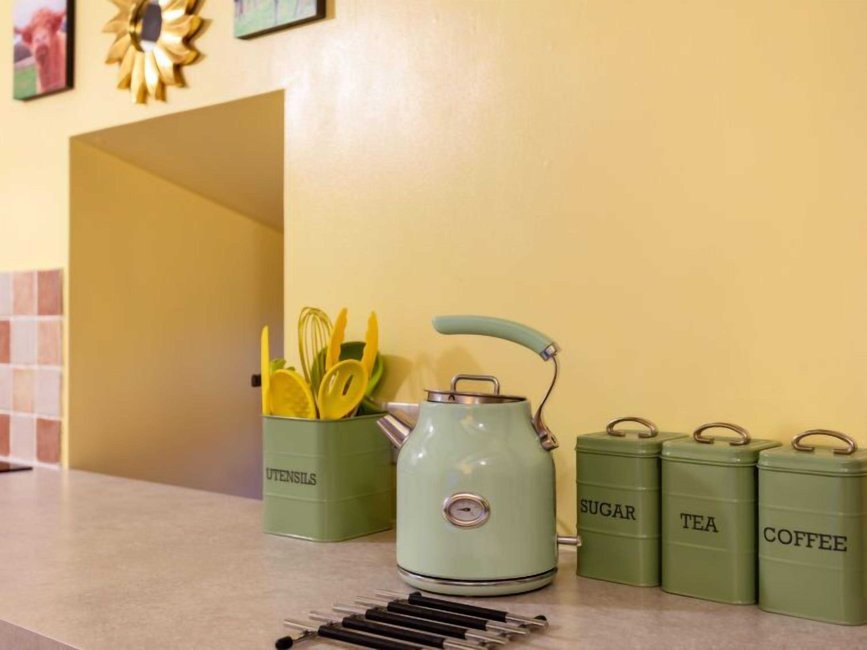 A kitchen counter with a green teapot, a utensil holder, and labeled tins for sugar, tea, and coffee.