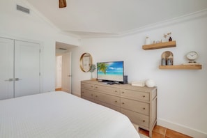 A bedroom with a bed, a TV on a dresser, a door, and a wall-mounted shelf with decorative items.