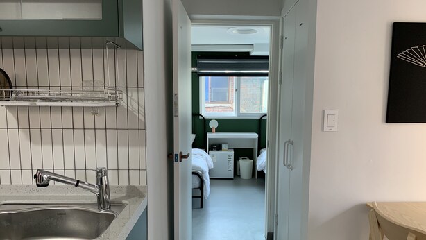 A narrow hallway with a kitchen area on the left, a bed with a nightstand in the center, and a small wooden table on the right. - Just Mansion (Seoul)