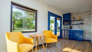 A modern kitchen with blue cabinets and a colorful backsplash, a small dining area with yellow chairs and a round table, and a large window offering a view of trees. - Groovy!!! at Odonata Escape -- retro chic container room in the country (Fredericksburg)
