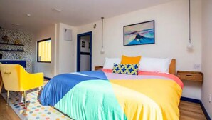 A bedroom with a colorful bedspread, a yellow chair, a wooden nightstand, and a painting on the wall. - Groovy!!! at Odonata Escape -- retro chic container room in the country (Fredericksburg)