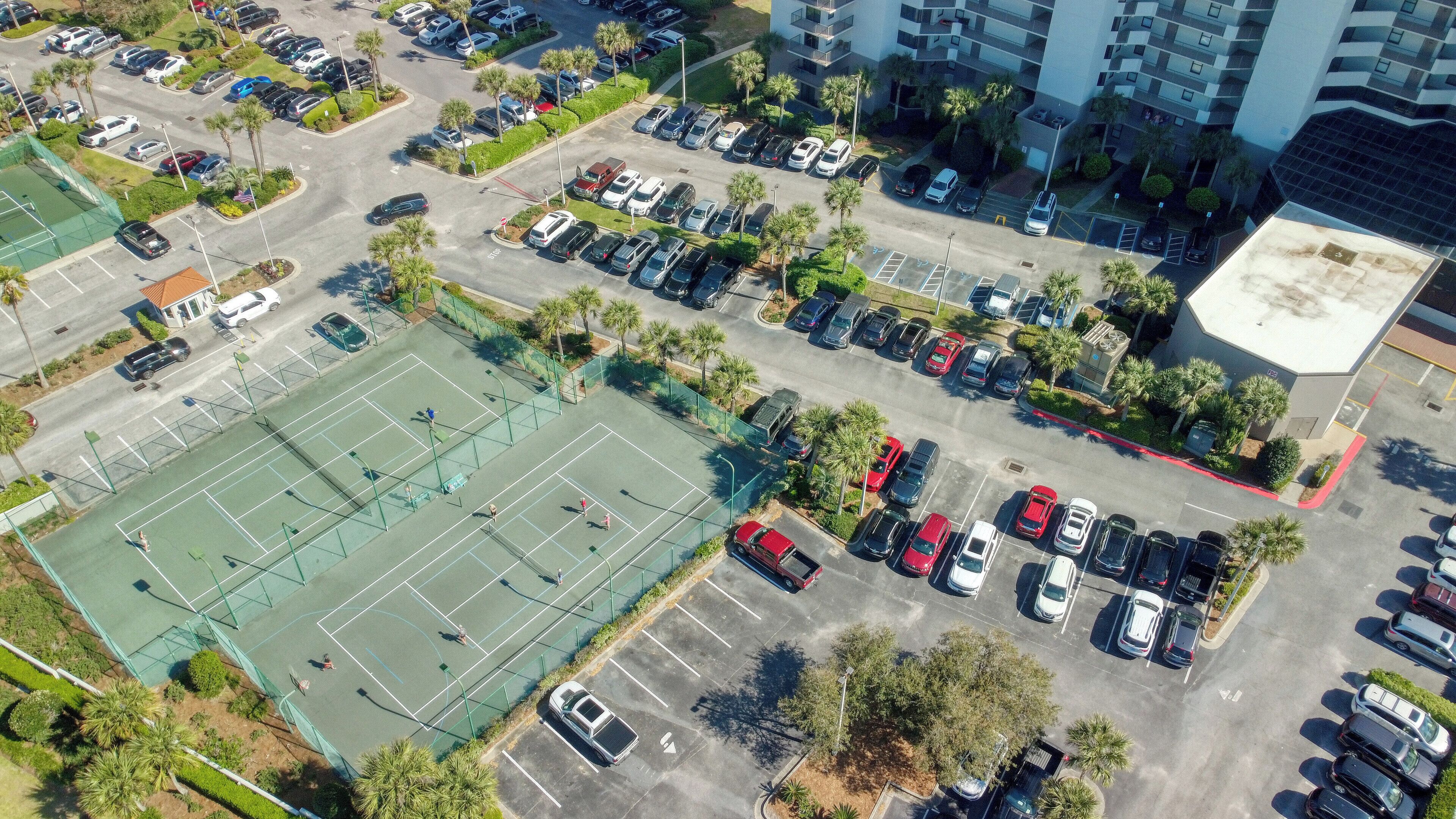 An aerial view of a parking lot with multiple rows of cars, a tennis court, and surrounding greenery.