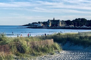 Una playa con un edificio histórico al fondo, un sendero arenoso que conduce al agua, y gente nadando y paseando por la orilla.