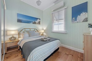 Une chambre à coucher comprenant un lit, des tables de chevet, une fenêtre avec des stores et des œuvres d’art murales.