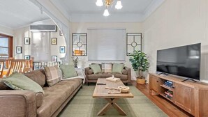 A living room with a brown sofa, a wooden coffee table, a TV on a wooden stand, and a large window with blinds. - Comfy Character QLD’r ~ 3Bed House w/Parking (Woolloongabba)
