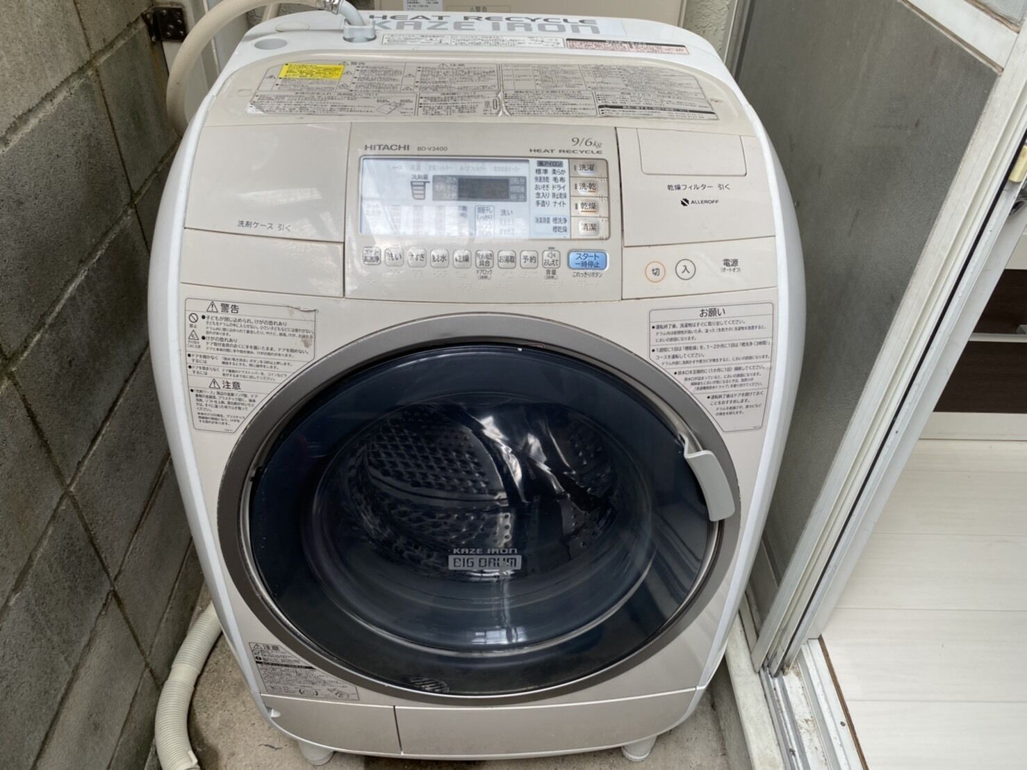 A front view of a white, front-loading washing machine with a digital display and control panel, located in a tiled room.