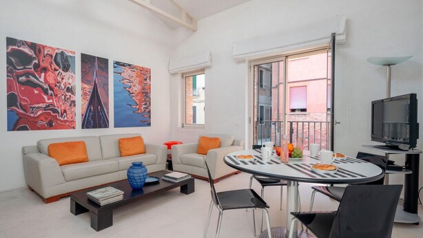 A modern living room with a sofa, coffee table, and a dining table with chairs. There are abstract paintings on the wall, a TV, and a window with a view of buildings. - The Waterview Alcove (Venice)