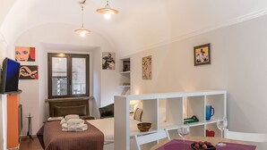 A hotel room with a bed, a dining table with two wine glasses, a white shelf unit, and artwork on the walls.