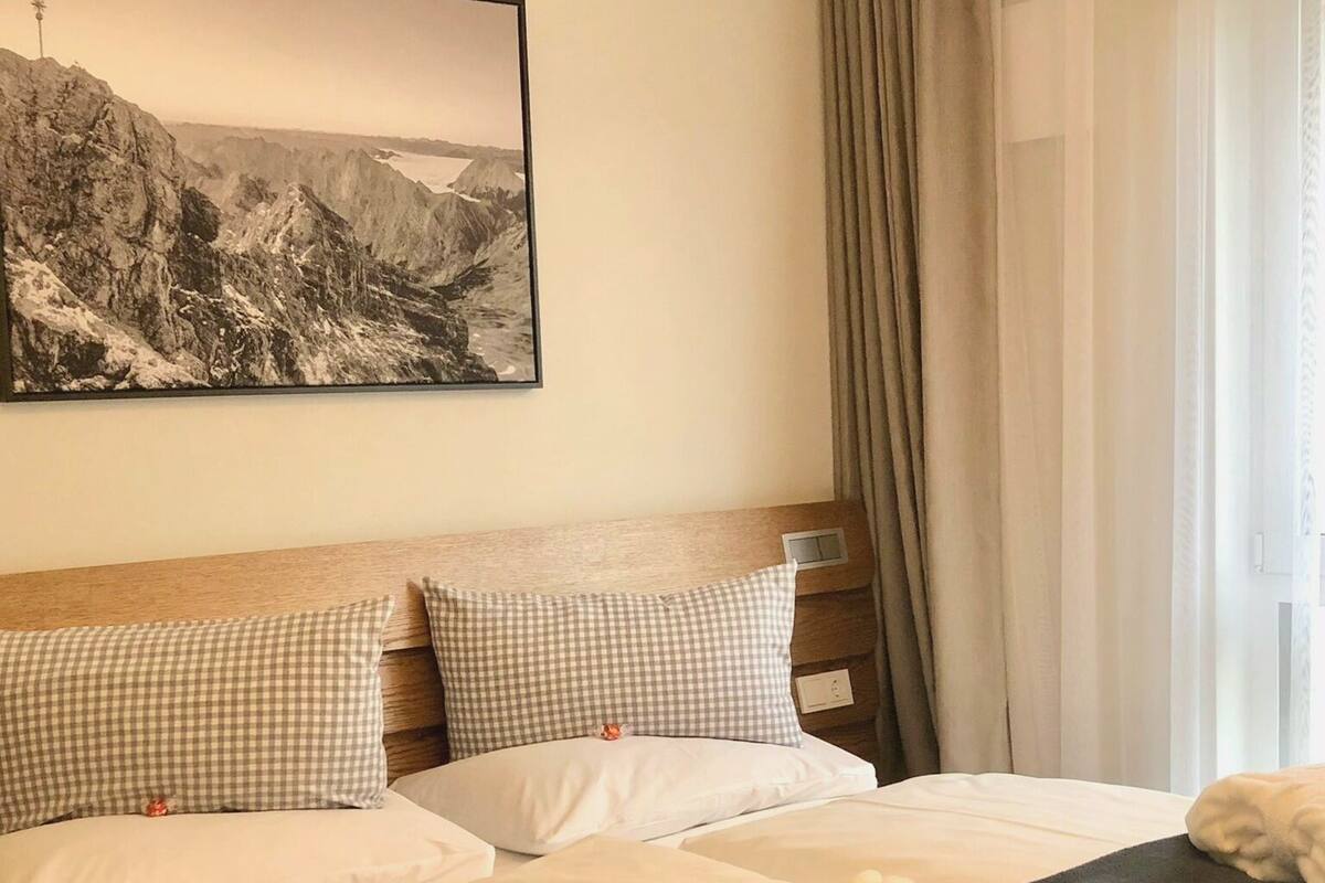 A hotel room with a bed, checkered pillows, a black blanket, a white robe, and a mountain landscape painting on the wall.