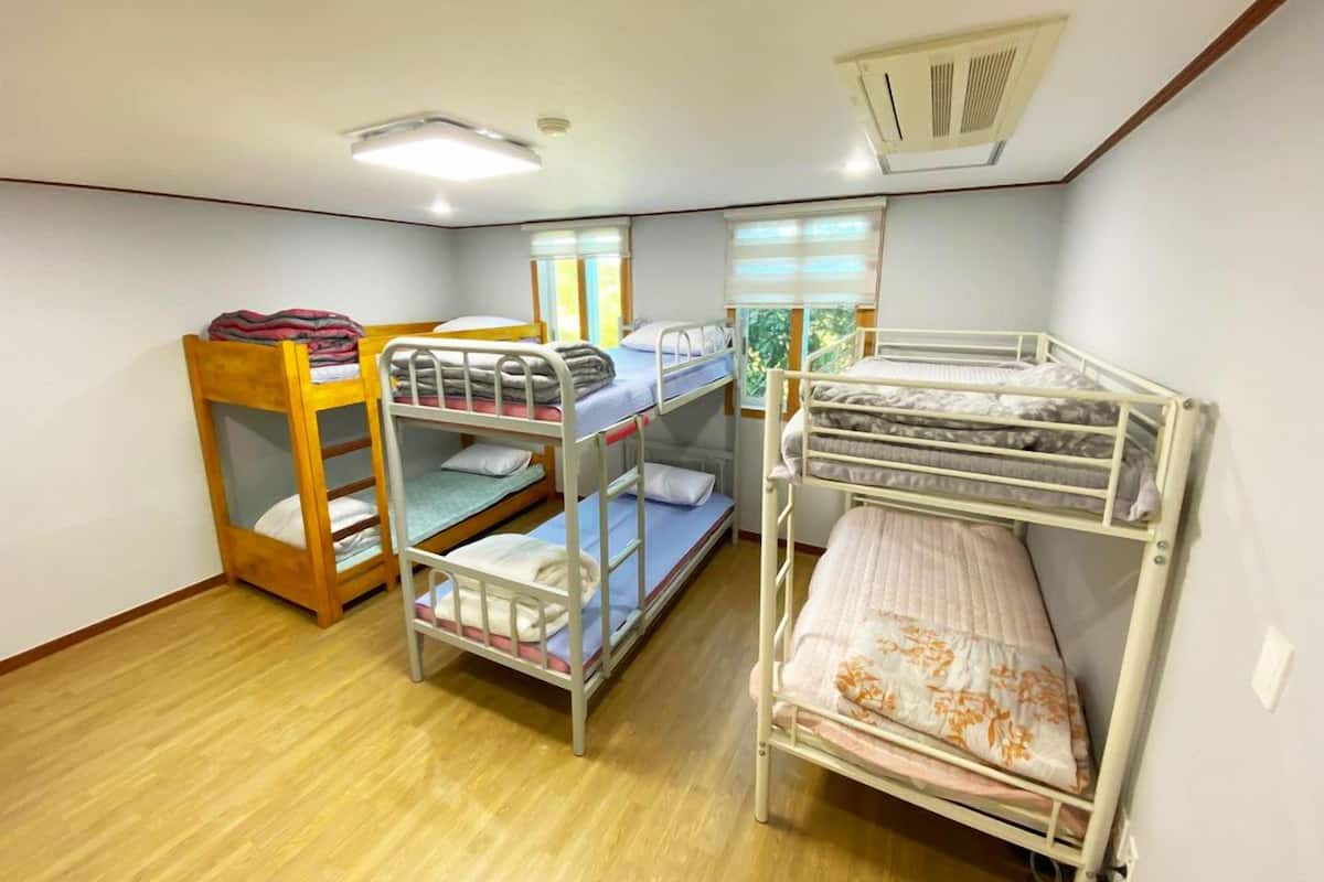 Une chambre de dortoir avec des lits superposés, une chaise en bois, une fenêtre avec des stores et une bouche d’aération au plafond.