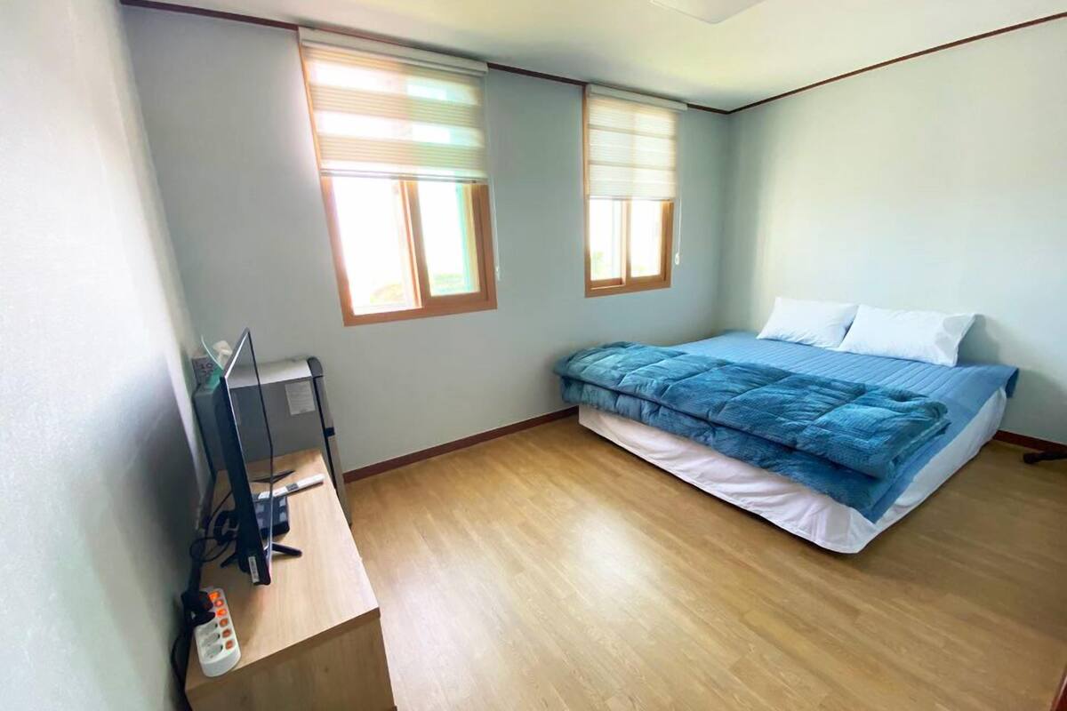 Une chambre à coucher avec un lit, deux fenêtres, un bureau avec un ordinateur et une chaise.