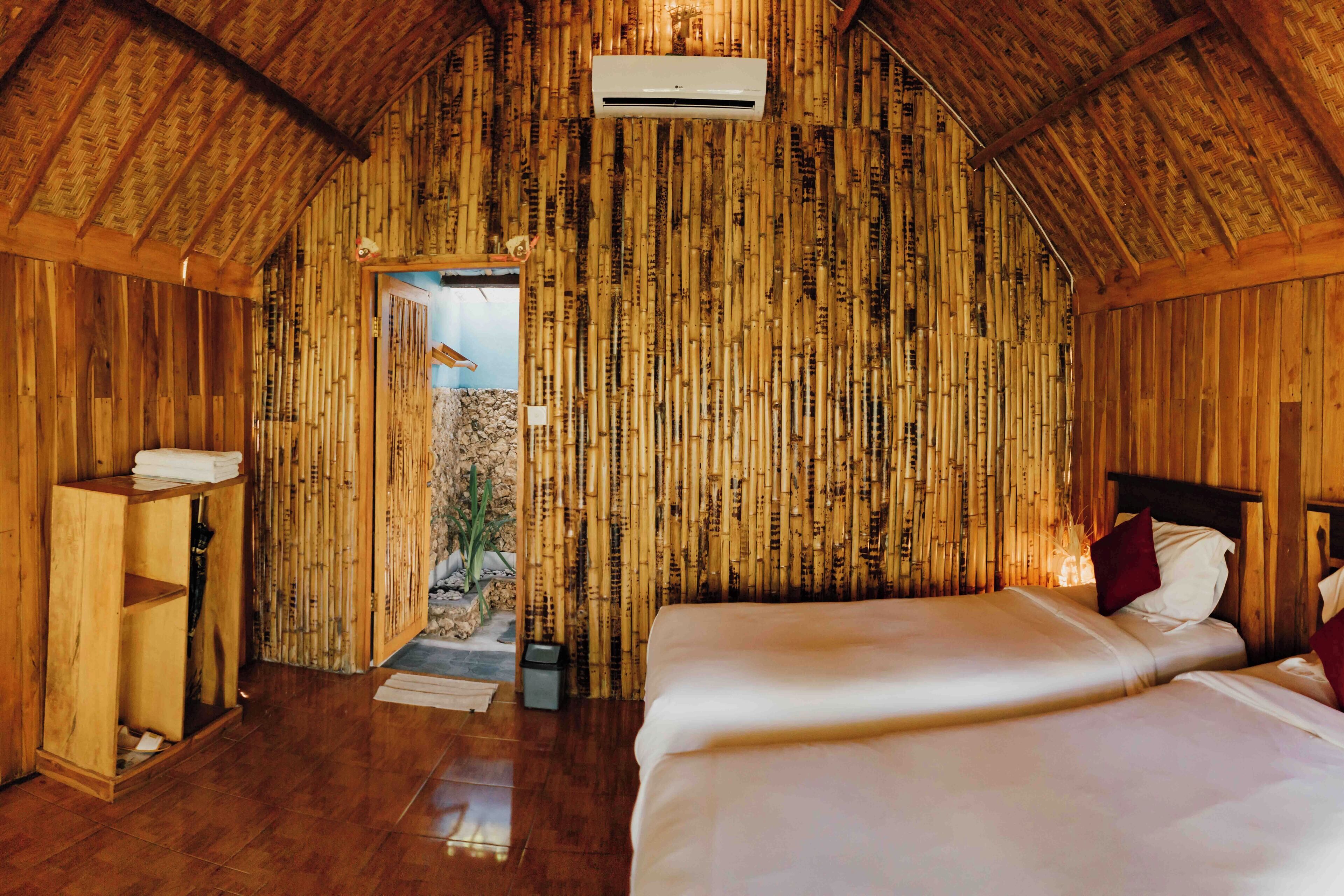 Quarto com paredes de bambu, teto de madeira e cama com roupa de cama branca e almofadas vermelhas.