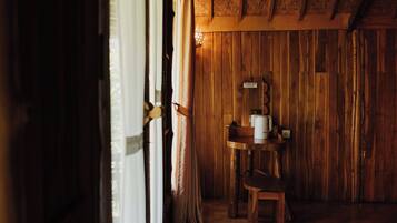 A rustic wooden room with a wooden table and chair, a small shelf with items, and a window with curtains.