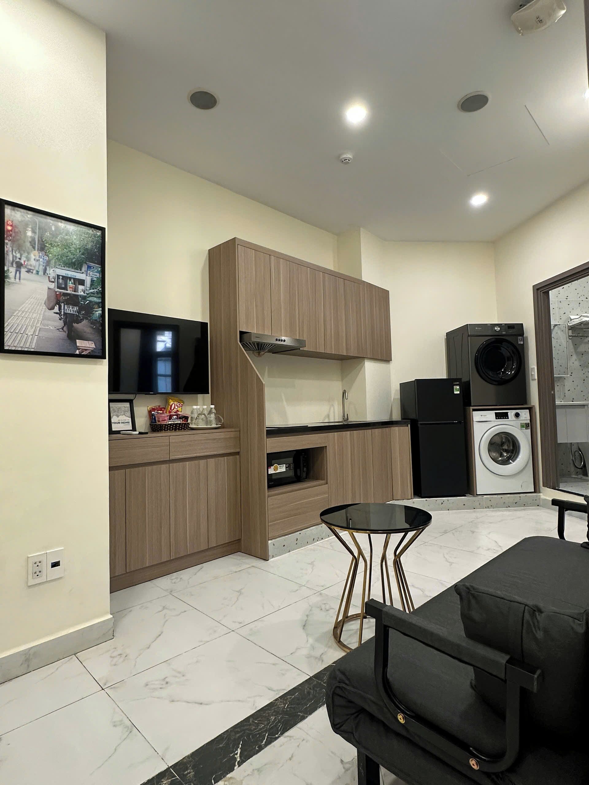 A compact kitchen with a built-in washing machine, a dining area with a black table and chair, and a mounted television.