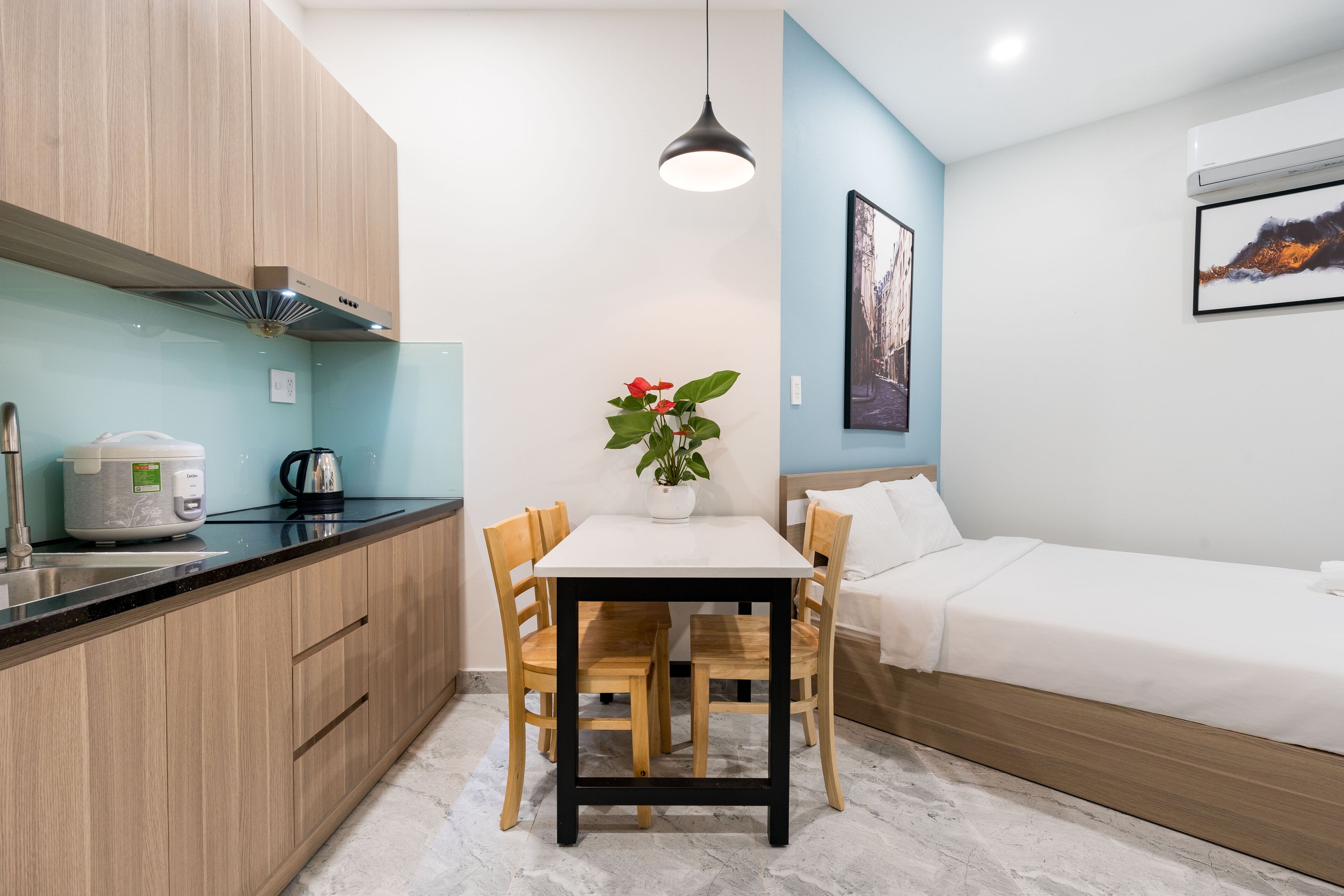 A compact kitchen with a dining table and chairs, a small potted plant, and a bed with a white mattress and pillows.