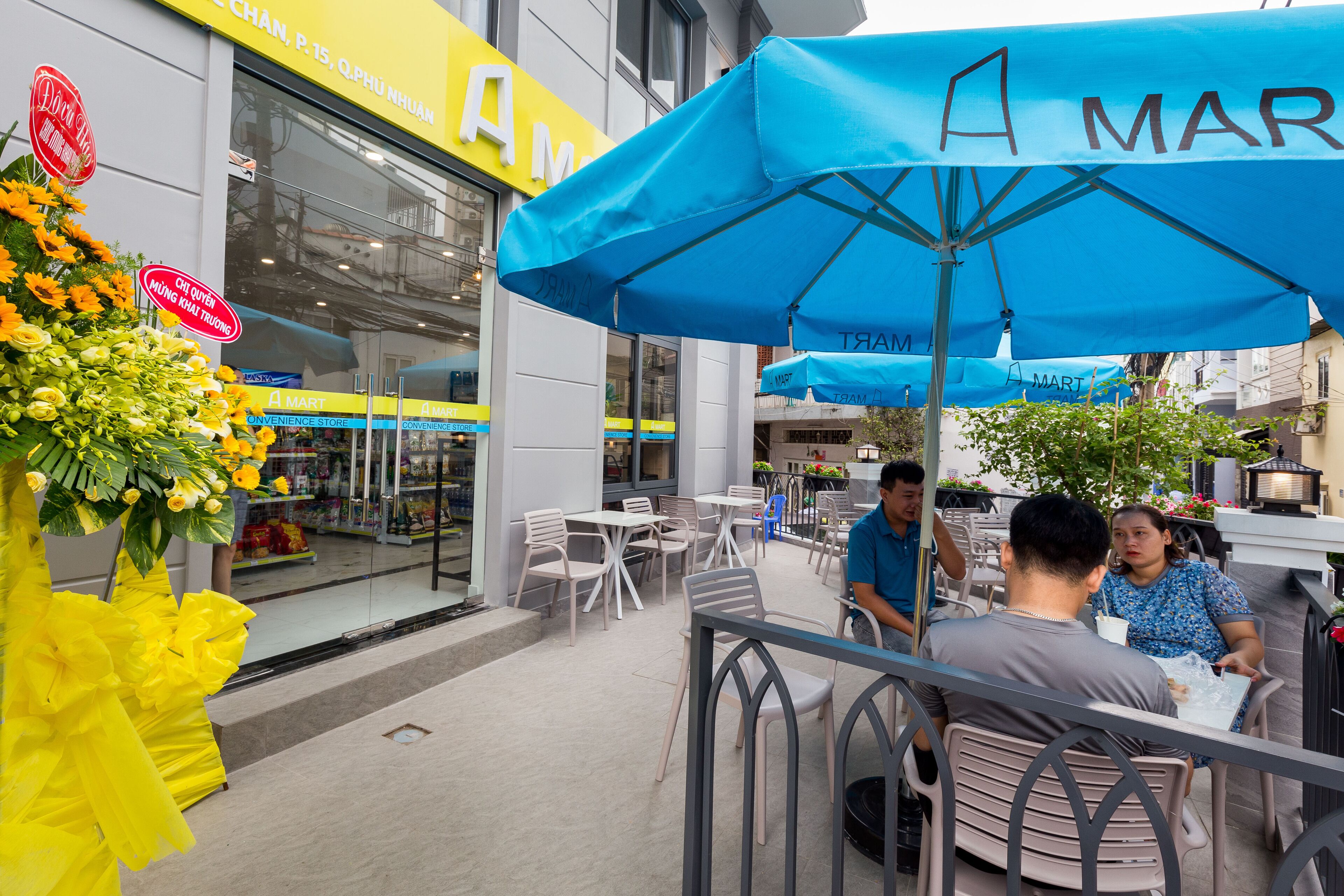 An outdoor café area with tables and chairs, a blue umbrella, and a storefront with a sign.