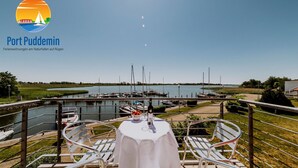 Un coin repas avec une table dressée pour deux personnes, donnant sur une marina où sont amarrés des bateaux et sur une étendue d’eau calme.