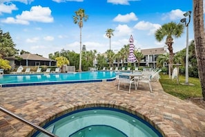 A swimming pool with lounge chairs and a small hot tub, surrounded by palm trees and a building with balconies.