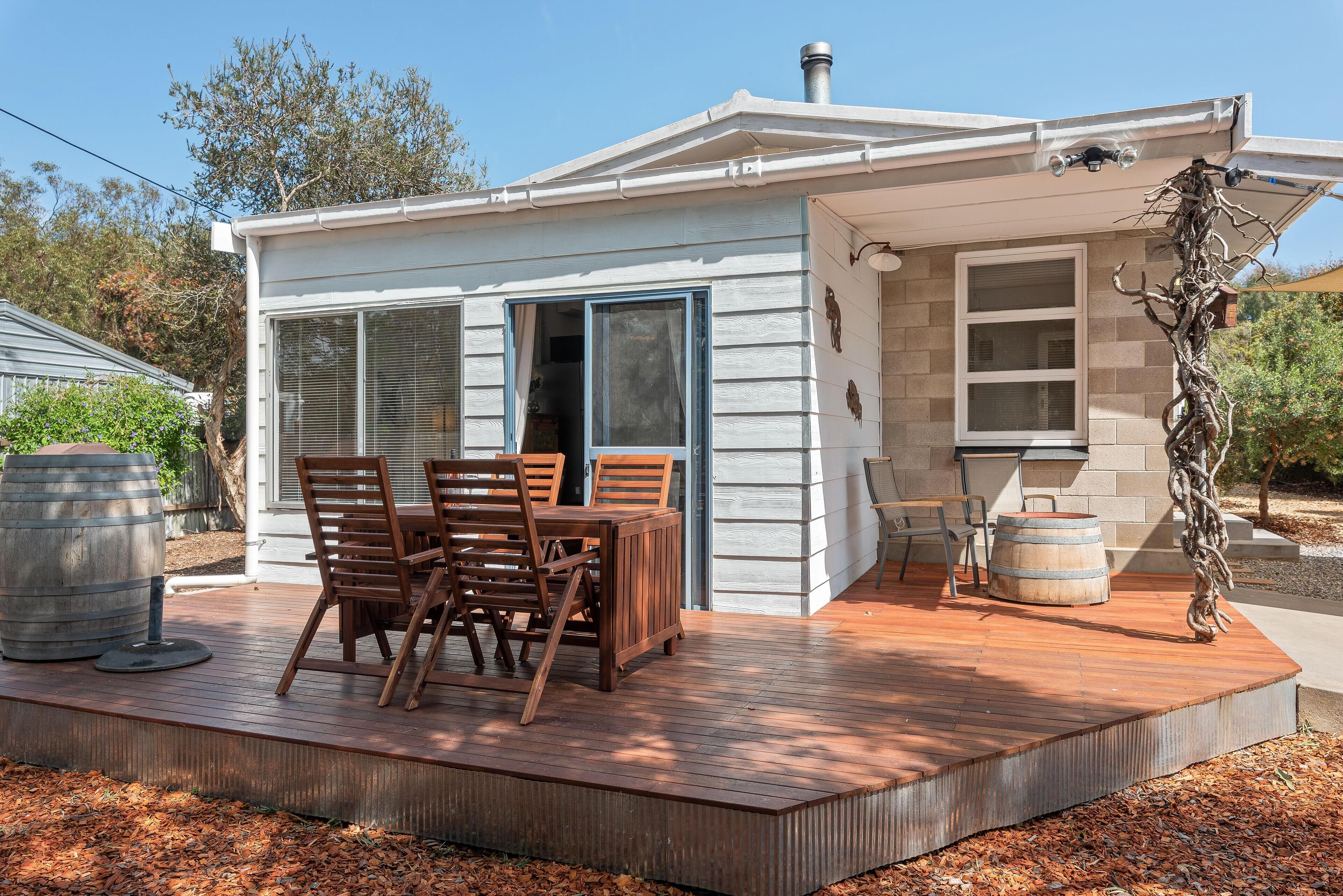 A small house with a deck, a dining table with chairs, and a large barrel.
