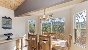 A rustic dining area with wooden furniture, a chandelier, and large windows offering a view of the forest.