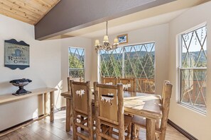Un comedor con mesa y sillas de madera, una lámpara de araña y ventanas grandes con vistas a árboles y montañas.