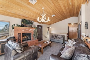 A cozy living room with a fireplace, leather sofas, a wooden ceiling, and a chandelier.