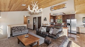 A spacious living area with a wooden ceiling, a large chandelier, a sofa set, a coffee table, and a kitchen in the background.