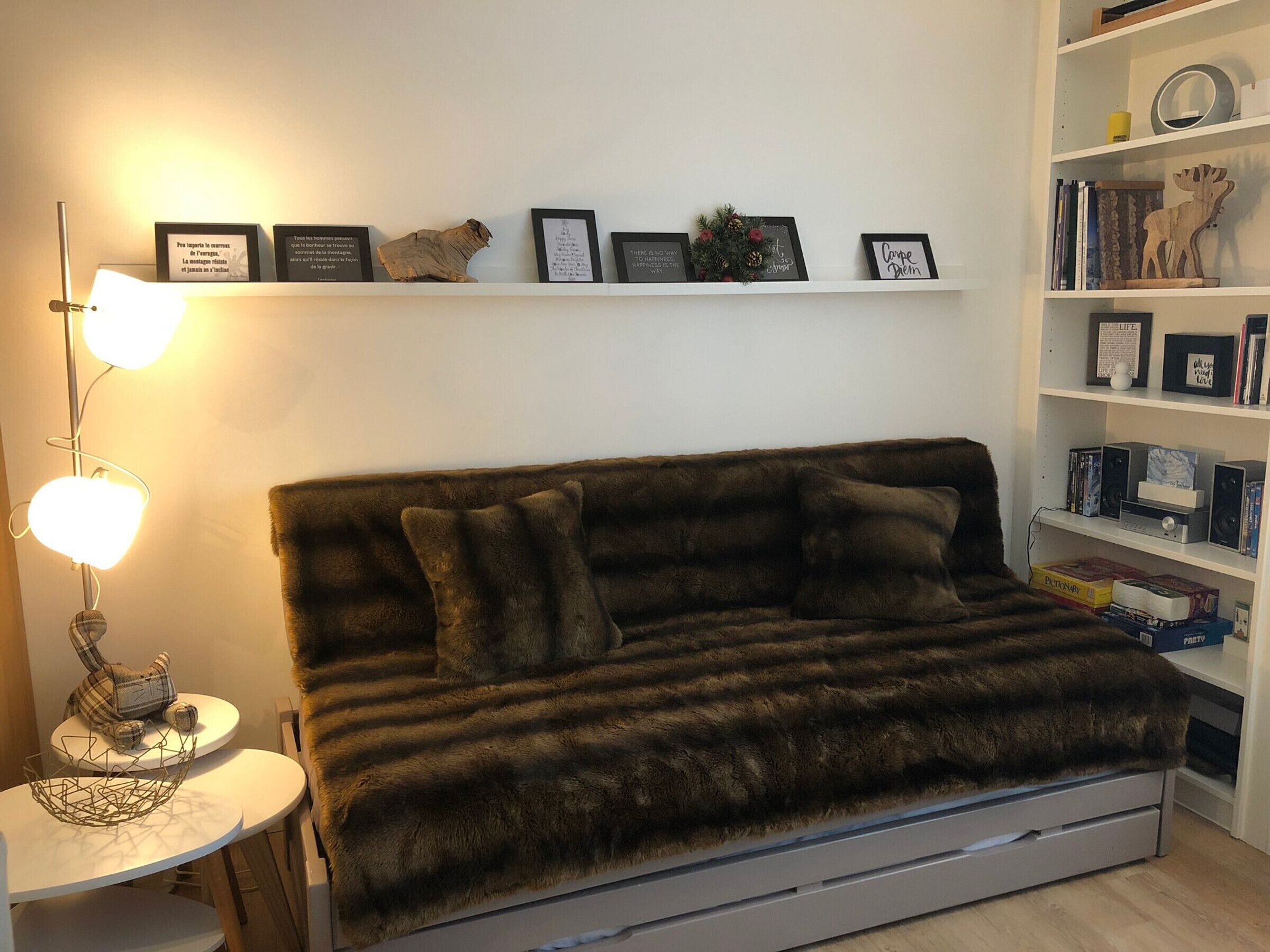 Ein gemütliches Wohnzimmer mit einem dunkelbraunen Sofa, einem kleinen runden Tisch mit Lampe sowie einem Bücherregal voller Bücher und Dekoration.