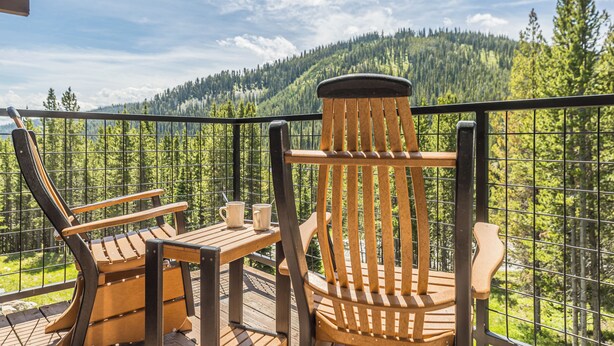 En träveranda med två Adirondack-stolar och ett bord, med utsikt över ett skogsbeklätt berg.
