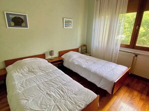 A room with two single beds, a wooden headboard, a nightstand, a lamp, and a window with curtains.