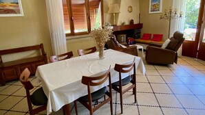 Uma sala de jantar com uma mesa posta para quatro pessoas, rodeada por cadeiras de madeira. Há um sofá com almofadas vermelhas ao fundo, uma lareira e uma janela com persianas.