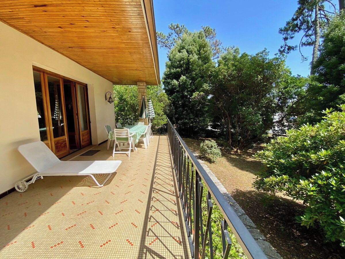 A balcony with a sun lounger and a table with chairs, surrounded by greenery and trees.